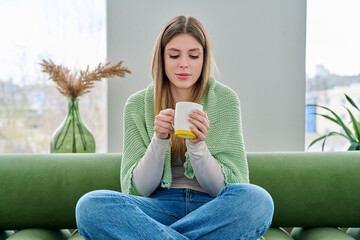 Portrait of relaxed beautiful young woman sitting at home on sofa with cup