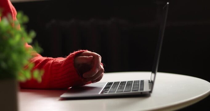 Close-up of laptop on table with hand in red sweater, taking a black  coffee cup onto table surface abd typing on keyboard, blurred background
