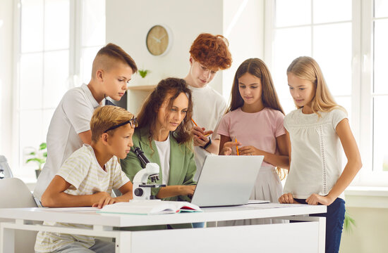 Group of school students and classmates sitting at the desk with microscope in classroom with young woman teacher and looking at the laptop monitor screen during a lesson. Education, science concept.