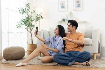 Fototapeta premium Happy Asian couple taking selfie at home, woman holding smartphone while man waves and smiles, sitting together in cozy living room, modern lifestyle, love, relationship, romance, and togetherness.