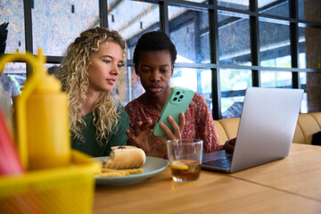 Diverse women friends sharing smartphone and laptop at lunch