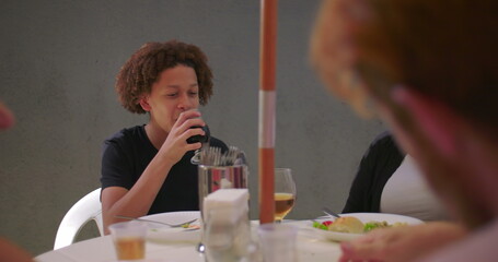 Young boy drinking from a glass and smiling, seated at an outdoor table during a family gathering...
