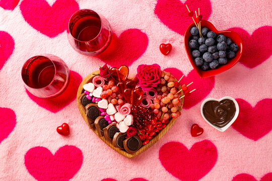 Snack board in shape of heart with berries, chocolate and sweets for Valentine's Day and two glasses of red wine on a pink background with hearts