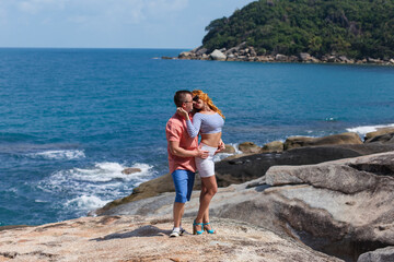 A young couple stands close on a sunlit granite rock outcrop overlooking a deep blue tropical bay, with a lush, green-covered hill in the background. The man wears an orange shirt and the woman has re