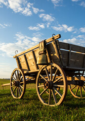 Obraz premium An old wooden wagon with large wheels sits in a grassy field under a blue sky with scattered clouds, featuring a hanging brass bell.