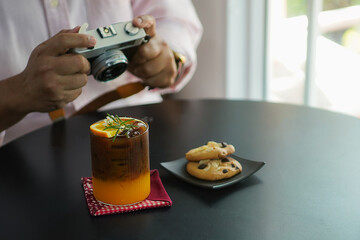 A refreshing iced coffee with orange juice garnish, decorated with rosemary and a small flower, served on a black table with cookies on a plate and a vintage camera in the background