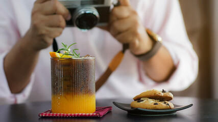 A refreshing iced coffee with orange juice garnish, decorated with rosemary and a small flower, served on a black table with cookies on a plate and a vintage camera in the background