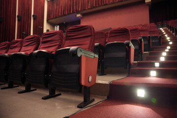 Rows of red theater chairs with black armrests with emergency lights on the stairs as a guide in the dark room.