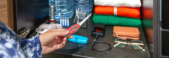 Unrecognizable woman hand holding multitool knife near emergency pantry closet with water, clothes, flashlight, batteries and powerbank. Female preparing 72 hour survival kit for disasters or war