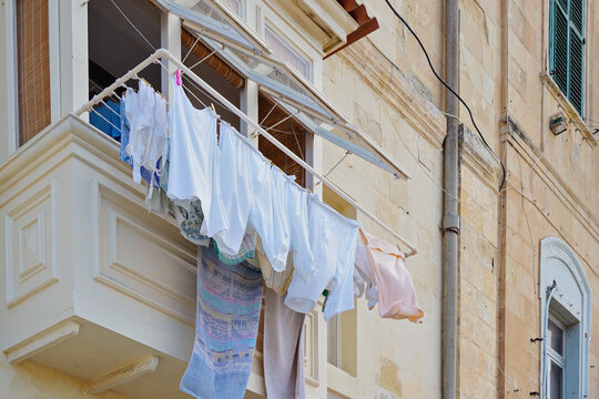 Sunlit Maltese balcony with clothesline, white fabrics drying above narrow street, warm limestone walls and colorful shutters, everyday Mediterranean scene