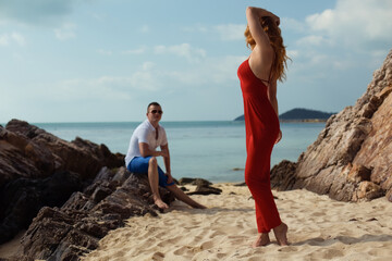 Description (199 characters):
A young woman with red hair and a fit build poses in a red jumpsuit with her back to the camera on a sandy beach. A man sits on a rock in the blurred background