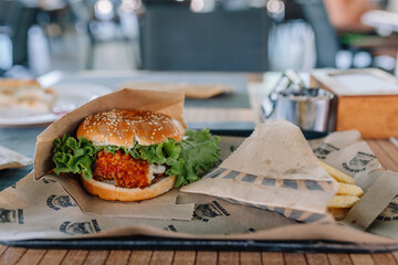 Close-up of a crispy chicken burger with fresh lettuce and sesame bun, served with French fries in a restaurant setting, wrapped in eco-friendly paper.