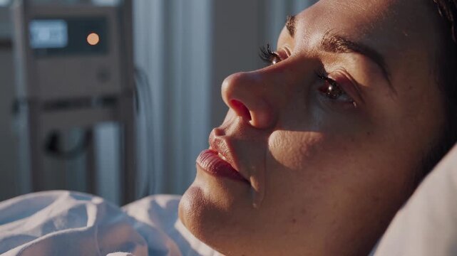 Close-up video shot of a woman lying in a hospital bed, capturing her thoughtful expression from a side angle in soft, natural lighting.