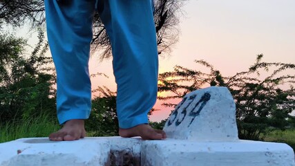 Barefoot man standing on a white milestone wearing blue pants with a sunset background