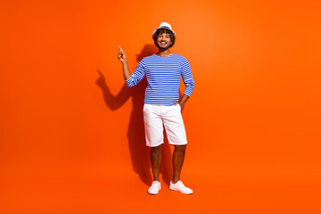 Cheerful young man in a striped sailor outfit standing against orange background pointing to the...
