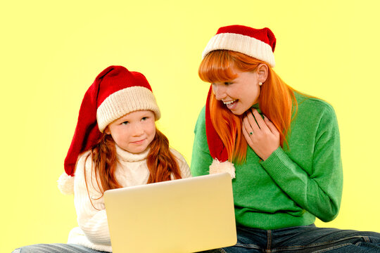 Mother and daughter with red hair wear Santa hats and use laptop in a Christmas holiday setting on a bright yellow background - Powered by Adobe