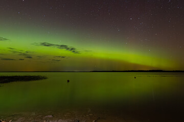 Bright green and red northern lights dance over a calm lake, casting vivid reflections on the water under a star-filled sky during a clear night.