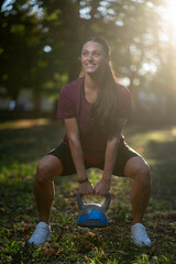 Fit woman performing kettlebell squat outdoors in park at sunrise, athletic posture, dedication, blurred background