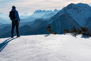 A hiker stands on a snowy ridge, gazing toward the dramatic peaks of Mittagskogel in the Karawanks, Bärentaler Kotschna, Carinthia, Austria, with layers of mountains fading into Slovenia.