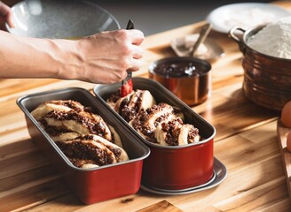 A person is shown brushing a glaze over rising Cozonac dough in baking tins. The Babka bread preparation is in progress, with ingredients on the wooden table nearby