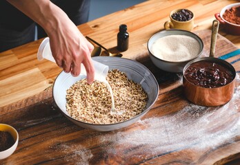 A person is pouring milk into a bowl of nuts, preparing the filling for a homemade cozonac on a floured wooden table, alongside other ingredients like sugar and melted chocolate