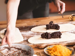 A person is carefully placing a rich, dark poppy seed filling onto flattened dough in preparation for baking a homemade Cozonac. Wooden table and ingredients are in view