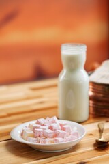 Cubes of Turkish delight are arranged on a white plate, sitting on a wooden table. A bottle of milk stands nearby, hinting at a traditional Eastern European breakfast