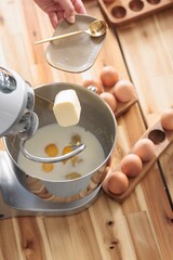 A stand mixer blends milk and eggs as butter is added. A wooden table is adorned with eggs and baking tools, setting the scene for making cozonac, maybe for Easter