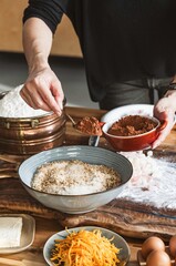 A person is making Cozonac. A spoonful of cocoa is being added to a bowl of dough. Ingredients like flour, eggs, cheese, and candied orange peels are visible