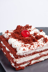 Red velvet cake slice with cream layers, white frosting, crumbs, and cherry on rectangular plate over dark surface, top view food photography