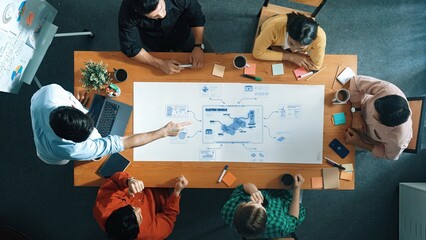 A top-down view of a diverse group of professionals engaged in a collaborative meeting, discussing innovative strategies with digital designs on the table in a modern office setting. Raster