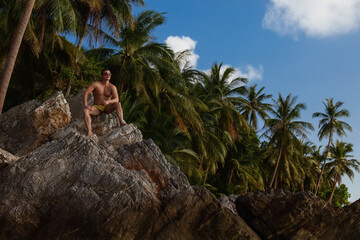 Muscular man in trunks sits perched on a large boulder at a tropical beach, looking up toward the light, with dense palm trees in the background