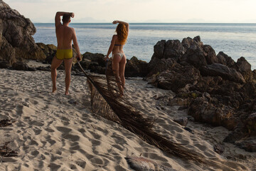 Fit couple posing on a sandy beach at dusk, with rock formations and the ocean in the background, a large dried palm leaf lies in the foreground.