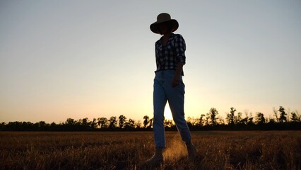 Adult agronomist in straw hat walking among barley plantation at dusk. Female farmer going through wheat meadow at sunset. Beautiful scenic landscape. Concept of agricultural business. Slow motion