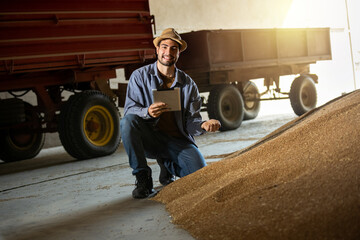 Young man holds soybeans in one hand and tablet in the other in warehouse, concept of modern agriculture, crop management, farming, logistics, food industry.