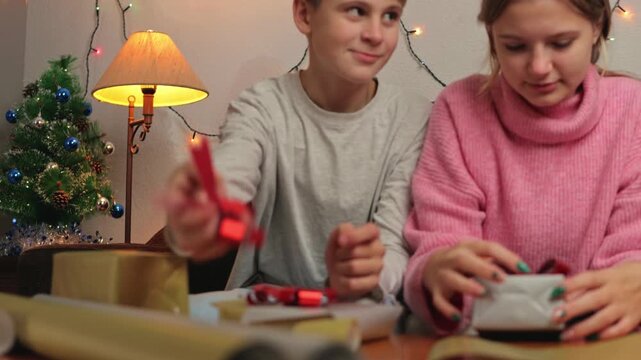 Cheerful siblings preparing for christmas eve, carefully wrapping gifts and decorating them with red ribbons in cozy room with decorated tree and festive lights for holiday season - Powered by Adobe