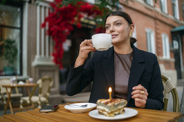 Joyful woman at an outdoor cafe celebrating with a slice of cake featuring a single birthday candle. She wears a smart black blazer and has a cup of tea on the wooden table.