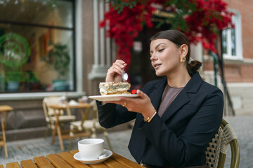 Joyful woman at an outdoor cafe celebrating with a slice of cake featuring a single birthday candle. She wears a smart black blazer and has a cup of tea on the wooden table.