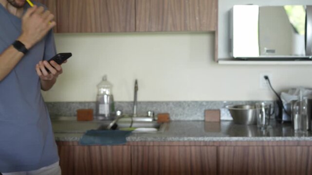 A young man in a kitchen brushes his teeth while engrossed in his phone, showcasing a modern blend of multitasking and phone addiction. A relatable moment reflecting technology's role in daily