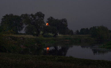 Red moon rising over river with reflection at night