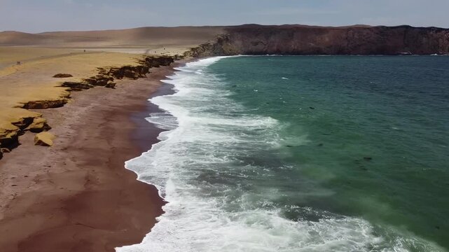 Drone footage flying over Playa Roja in Paracas, Peru, featuring the striking red sands, dunes, and the Pacific Ocean. The remote coastal desert offers a serene and dramatic landscape, captured in a b