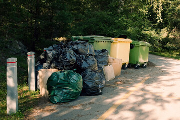 Large pile of garbage bags and cardboard boxes near outdoor trash bins in a green park area. Waste management and environmental pollution problem.