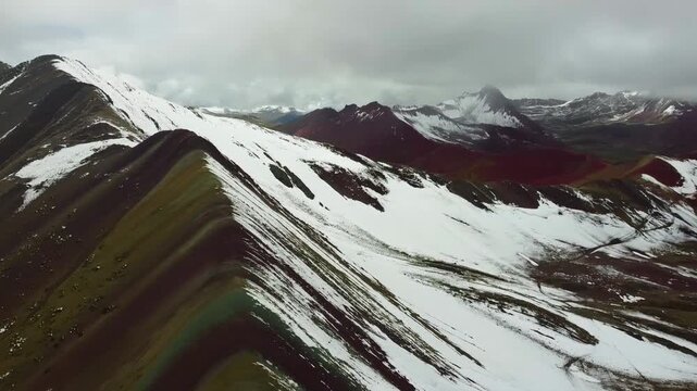 Aerial drone footage of Rainbow Mountain in the Peruvian Andes, Vinicunca, showing colorful striped peaks partly covered with snow and the surrounding vibrant mountains.