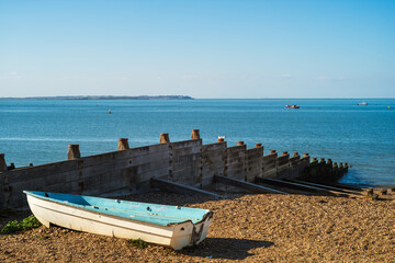 View across to the Isle of Sheppey from Whitstable beach. A wooden groyne and small blue boat are on the pebble beach.