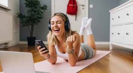 Happy woman laughing while watching something on her phone with headphones on, lying on yoga mat