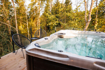 A modern hot tub on a wooden deck surrounded by tall trees, with glass railings, two black chairs, a small table, and warm lighting in a forest setting.