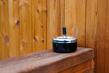 A metal ashtray with a black base and blue push button sits on a wooden surface, surrounded by vertical wooden panels with a natural finish.