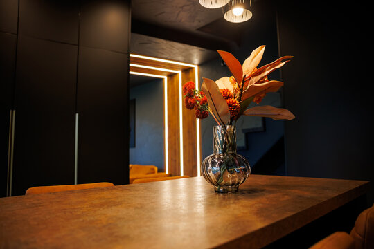 A sleek wooden table with a glass vase of orange and red flowers sits in a modern room with dark walls, warm lighting, and illuminated wooden panels. - Powered by Adobe