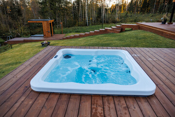 Modern hot tub on a wooden deck with bubbling water, surrounded by grass, tall trees, string lights, and a nearby wooden structure.