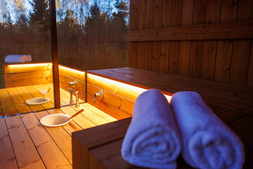A wooden sauna interior with warm lighting, polished surfaces, white towels, a ladle, and a water basin. A glass window reveals trees outside.